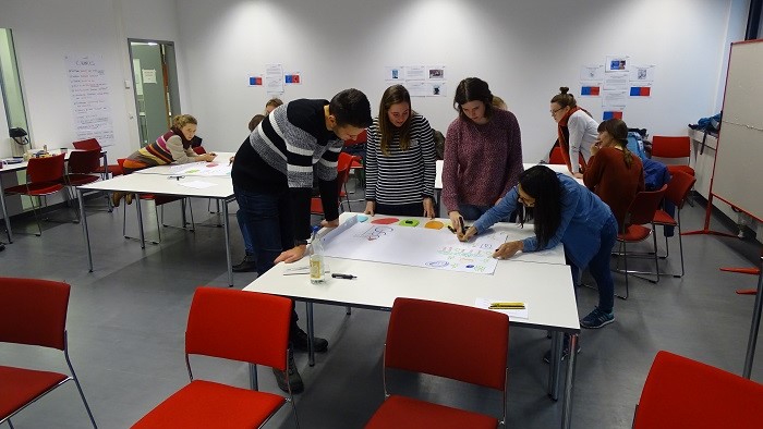 An drei Tischen verteilt sitzen oder stehen Studierende. Vor ihnen liegen leere Poster, auf die sie schreiben oder malen.