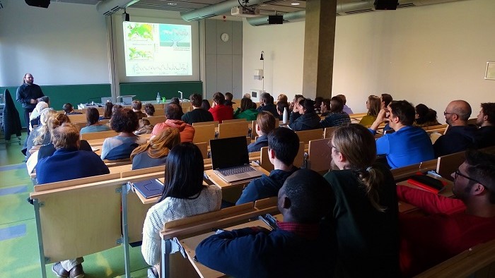 In einem Hörsaal sitzen viele Zuhörer, die konzentriert nach vorne schauen. Vorne steht Prof. Dr. Manuel Steinbauer und hält seinen Vortrag.