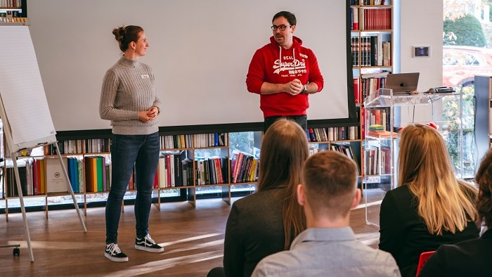 Two speakers stand before a group of young people.