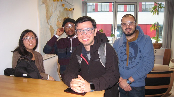 Vier junge Menschen blicken in die Fotokamera und setzen sich währenddessen eine Schutzbrille auf.