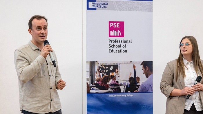Das Bild zeigt zwei Personen mit Mikrofonen neben einem Roll-up-Banner der Professional School of Education der Universität Würzburg.
