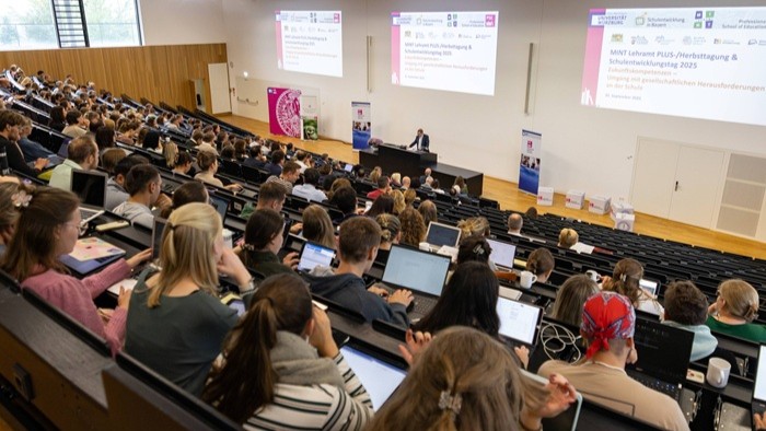 © Christoph Weiß Das Bild zeigt einen großen Hörsaal, in dem zahlreiche Teilnehmende einer Präsentation folgen, während auf zwei Leinwänden Informationen zur Veranstaltung angezeigt werden.