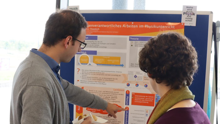 Das Bild zeigt zwei Personen, die vor einem Poster zum Thema „Eigenverantwortliches Arbeiten im Physikunterricht“ stehen und gemeinsam Inhalte besprechen.
