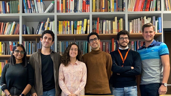 A group of six young people photographed in front of a library wall.