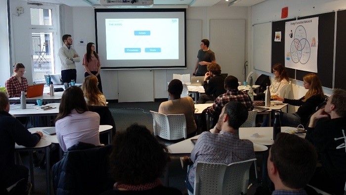 A group of young people are standing and sitting in a room with a screen.