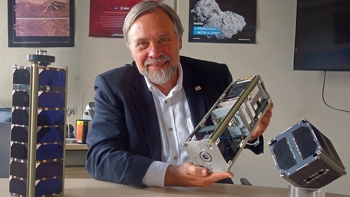 Klaus Schilling mit den an der Universität Würzburg gebauten Kleinsatelliten UWE und NetSat im Vordergrund sowie den Satelliten HUYGENS und ROSETTA aus seiner früheren Industrietätigkeit auf Postern.