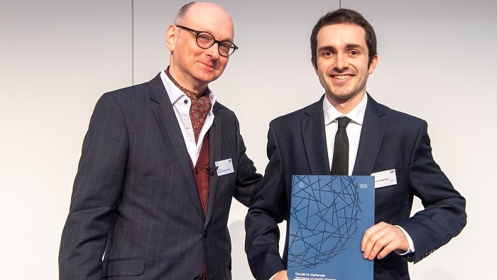 The Dean of the Department of Mathematics and the prize winner of the TopMath Award 2019 are stood in front of a grey wall in the lecture theatre. They are smiling. The awardee Johannes Bäumler holds a blue folder with the logo of the Department of Mathematics. 