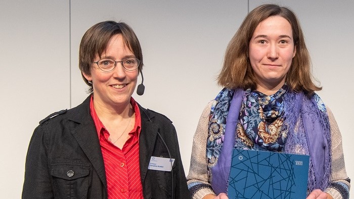 The professor and the prize winner are stood in front of a grey wall in the lecture theatre. The awardee Vera Pazukhina holds a blue folder with the logo of the Department of Mathematics.