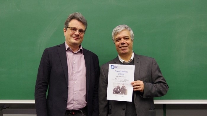 Two men are standing in front of a blackboard. The man on the right is holding a certificate. 