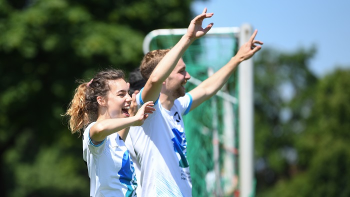 A young woman and a young man in soccer jerseys cheer on the sidelines of the soccer match.