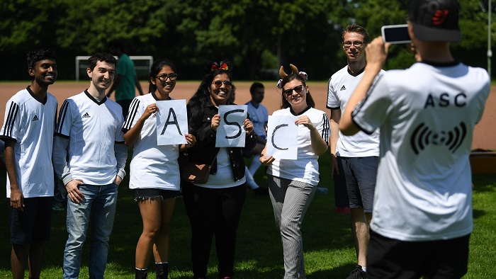 Eine Gruppe junger Menschen in Fußball-t-shirts stehen am Spielfeldrand und halten lächelnd Plakate mit den Buchstaben ASC in die Kamera.