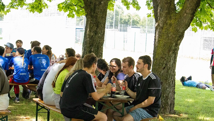 Studierende essen an Biertischen unter Bäumen.