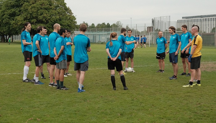 Eine Gruppe von jungen Männern und Frauen steht in Fußballtrikots auf dem Fußballplatz. Sie bilden einen Kreis um den Mannschaftskapitän, der etwas erklärt.