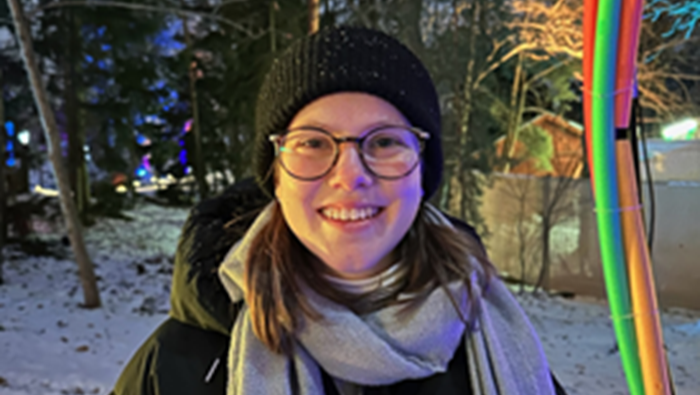 Das Bild zeigt eine Person mit braunen Haaren, die eine graue Jacke und einen Schal trägt, in einer winterlichen, festlich beleuchteten Außenumgebung. Im Hintergrund sind bunte Lichter und Tannenbäume zu sehen.