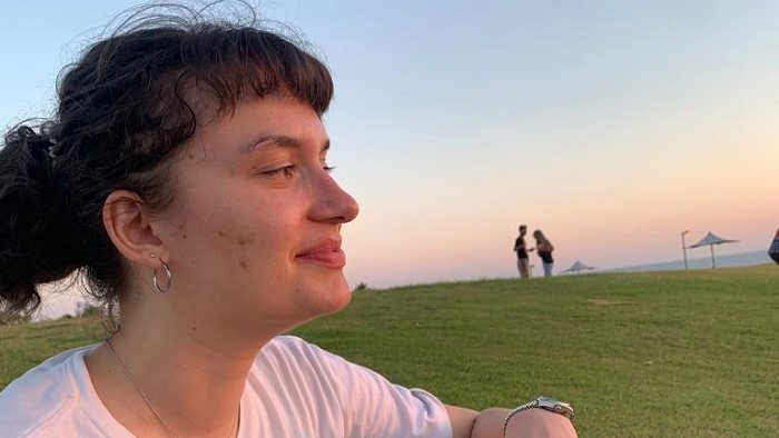 A young woman in sporty clothes sits on a meadow near the beach in front of a blue sky.