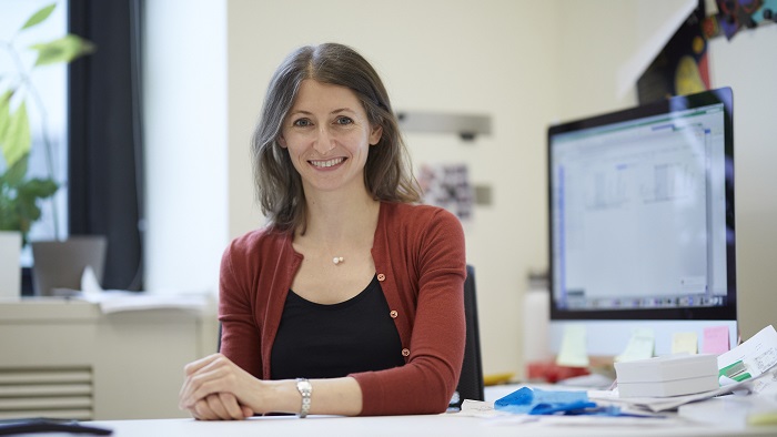 A woman with long, brown hair sits at her desk and smiles at the camera.