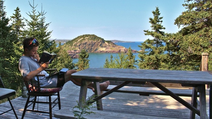 A person reading on a wooden terrace overlooking the sea and forested cliffs.