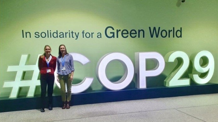 Two people are standing in front of a photo wall displaying the text "#COP29 In solidarity for a Green World".