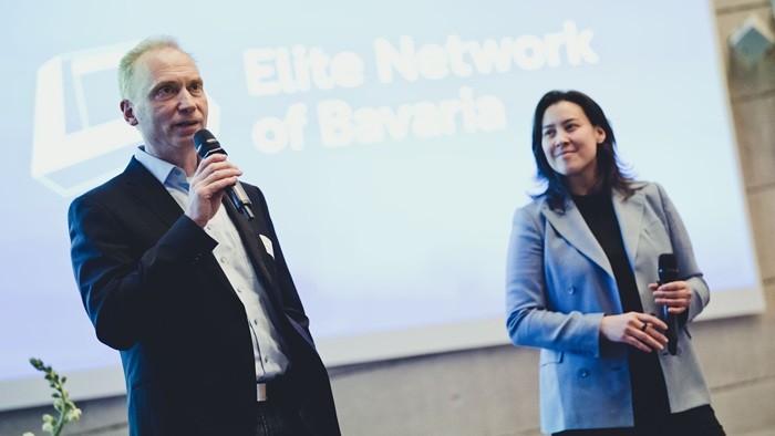 Zwei Personen stehen mit Mikrofon auf einer Bühne vor einer großen Präsentationsfläche des Elitenetzwerks Bayern.