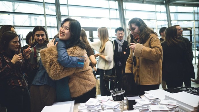 Teilnehmende begrüßen sich mit einer Umarmung vor dem Merchandise-Stand des Elitenetzwerks.