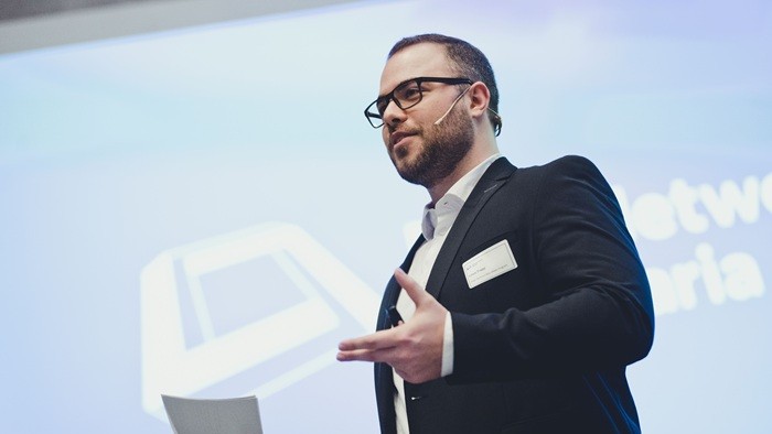 Ein Moderator spricht zum Publikum vor einer großen Präsentationsfläche.