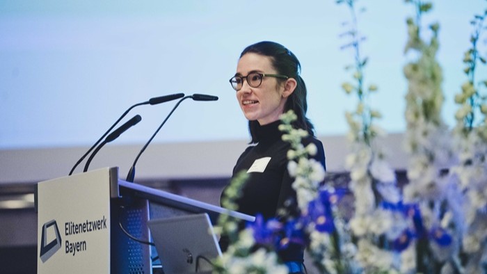 Eine Rednerin steht an einem Pult vor einer großen Präsentationsfläche.