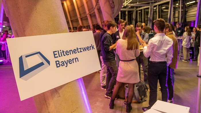 Eine Gruppe junger Menschen unterhält sich an einem Stehtisch; Neben Ihnen ist das neue Logo des Elitenetzwerks Bayern an einer Säule befestigt.