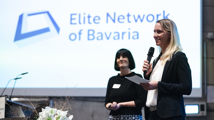 Two women are standing on stage and speak to the audience.