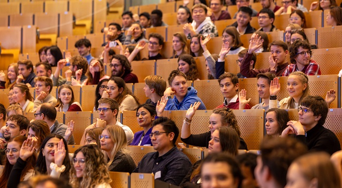 Numerous young men and women are sitting in the rows of the lecture hall. They laugh and some of them raise their hands.