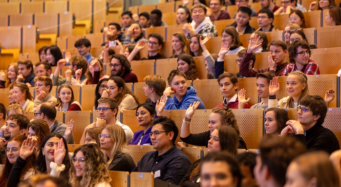 In den Reihen des Hörsaals sitzen zahlreiche junge Männer und Frauen. Die lachen und ein Teil von ihnen hebt die Hand.