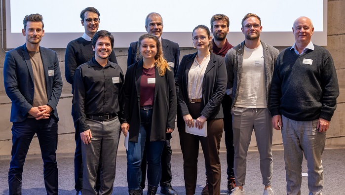 A group of nine men and women are standing on the stage of the lecture hall and smile into the camera.