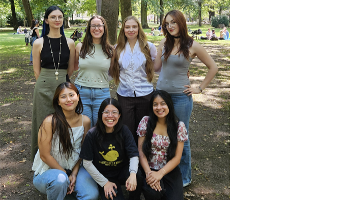Five people are standing next to each other in a green park with lots of trees, wearing casual clothes and posing for a photo.