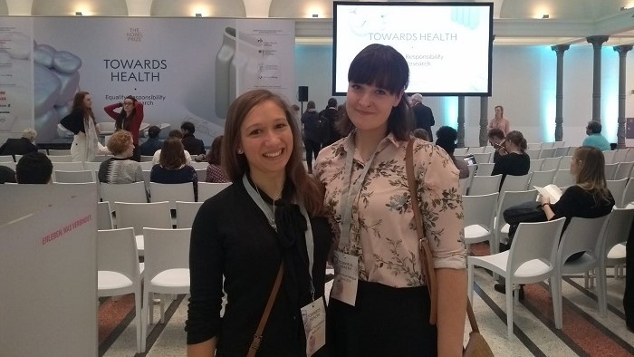 Zwei Studentinnen stehen vor einem Bildschirm, der das Motto des Nobel Prize Dialogue 2019 “Towa-rds Health – Equality, Responsibility and Research” zeigt.