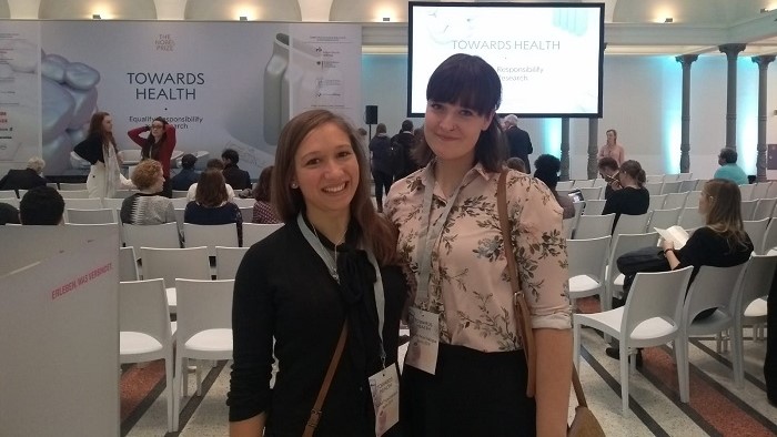 © Frederike Schäfer Zwei Studentinnen stehen vor einem Bildschirm, der das Motto des Nobel Prize Dialogue 2019 “Towa-rds Health – Equality, Responsibility and Research” zeigt.