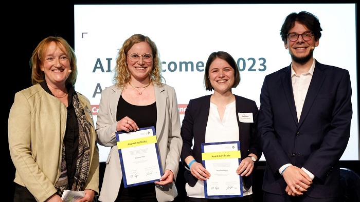 Four people standing side by side in front of a beamer projection. In the middle, two young females hold certificates in their hands.