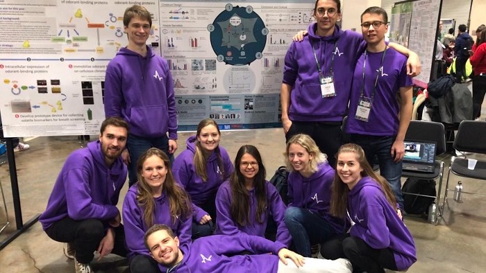 A group of people in purple sweaters in front of their poster. 
