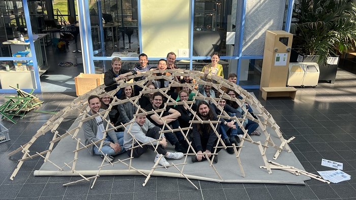 A group of young people squats under a construction made of wooden sticks.