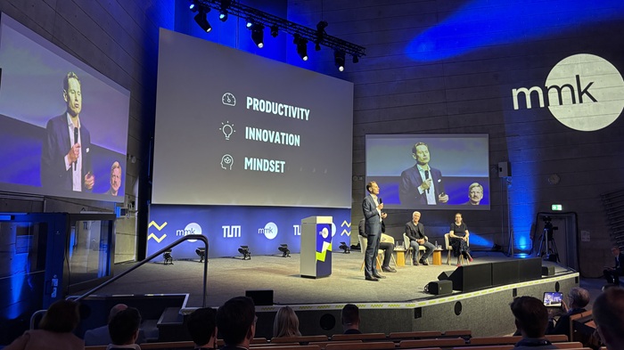 A speaker is giving a presentation on a large stage lit by blue lights.