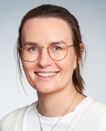 Porträtbild einer Frau mit zurückgebundenem braunem Haar, die ein weißes Oberteil und eine helle Strickjacke trägt, ergänzt durch eine Halskette.