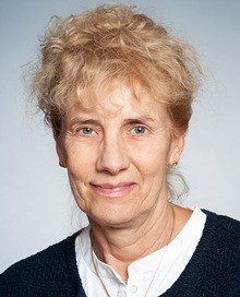 © Johannes Haas Porträtbild einer Frau mit lockigem, blondem Haar, die ein weißes Hemd unter einem dunklen Strickpullover trägt.