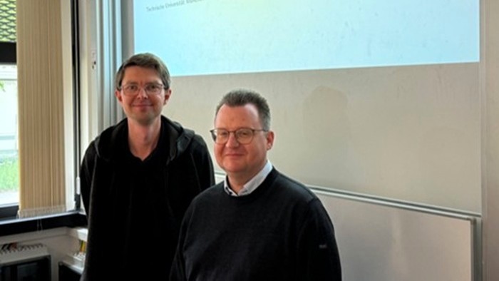 © Elite Graduate Program "Software Engineering" Two people are standing in a room in front of a white wall and a projection screen. In the background, there is a window with vertical blinds. Both people are wearing dark clothing, one standing slightly further forward, the other further back.