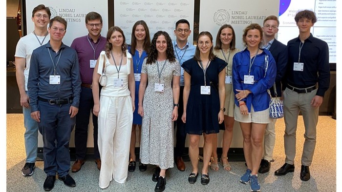 © Katia Werkmeister A group of people is standing in front of a wall with the Lindau Nobel Laureate Meetings logo and wearing name tags.