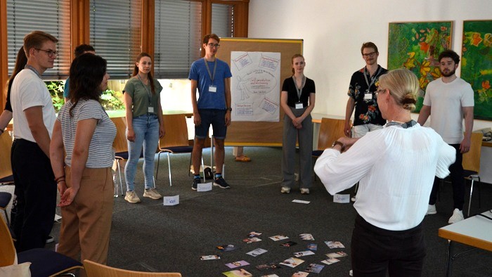 Personengruppe im Workshop-Kreis mit Karten auf dem Boden und Flipchart im Hintergrund.