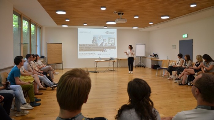 Ein Vortrag findet in einem Raum statt, in dem die Teilnehmenden im Kreis sitzen und einer Person zuhören, die vor einer Projektionsleinwand spricht.