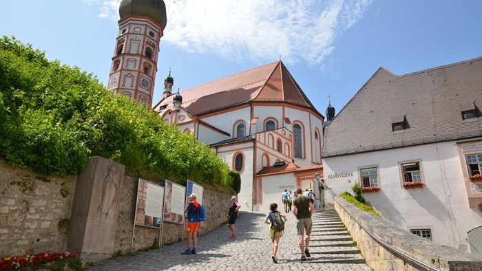 Eine Gruppe von Menschen geht einen Kopfsteinpflasterweg hinauf zu einer großen, reich verzierten Kirche mit rotem Dach und hohem Turm, umgeben von Grünflächen und weiteren Gebäuden.