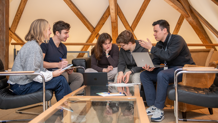 © Anita Gall / Max Weber-Programm Das Bild zeigt eine Gruppe von fünf Personen, die in einem modernen Raum mit Holzbalkenstruktur um einen Tisch sitzen und gemeinsam an Laptops und Notizen arbeiten.