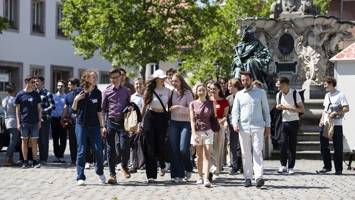 © Uwe Niklas / Max Weber Program A large group of young adults is walking together across a cobblestone square in front of a fountain and historic buildings.