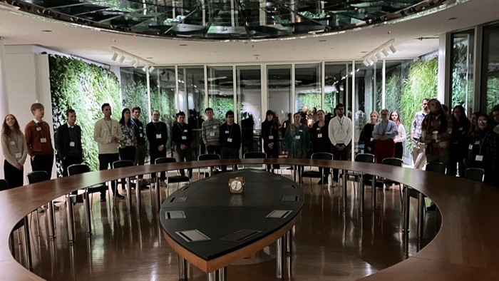 A group standing around a large oval table in a room with glass walls and greenery.