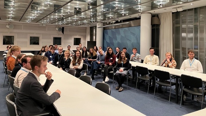 © Mylène Wienrank A large group of people seated in a modern conference room at long tables, facing the the front.