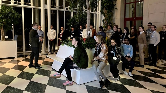 A group standing and sitting in a bright hall with plants, while one person speaks at the front.